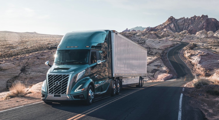 A teal semi-truck hauling a white trailer drives on a winding road through a rocky desert landscape